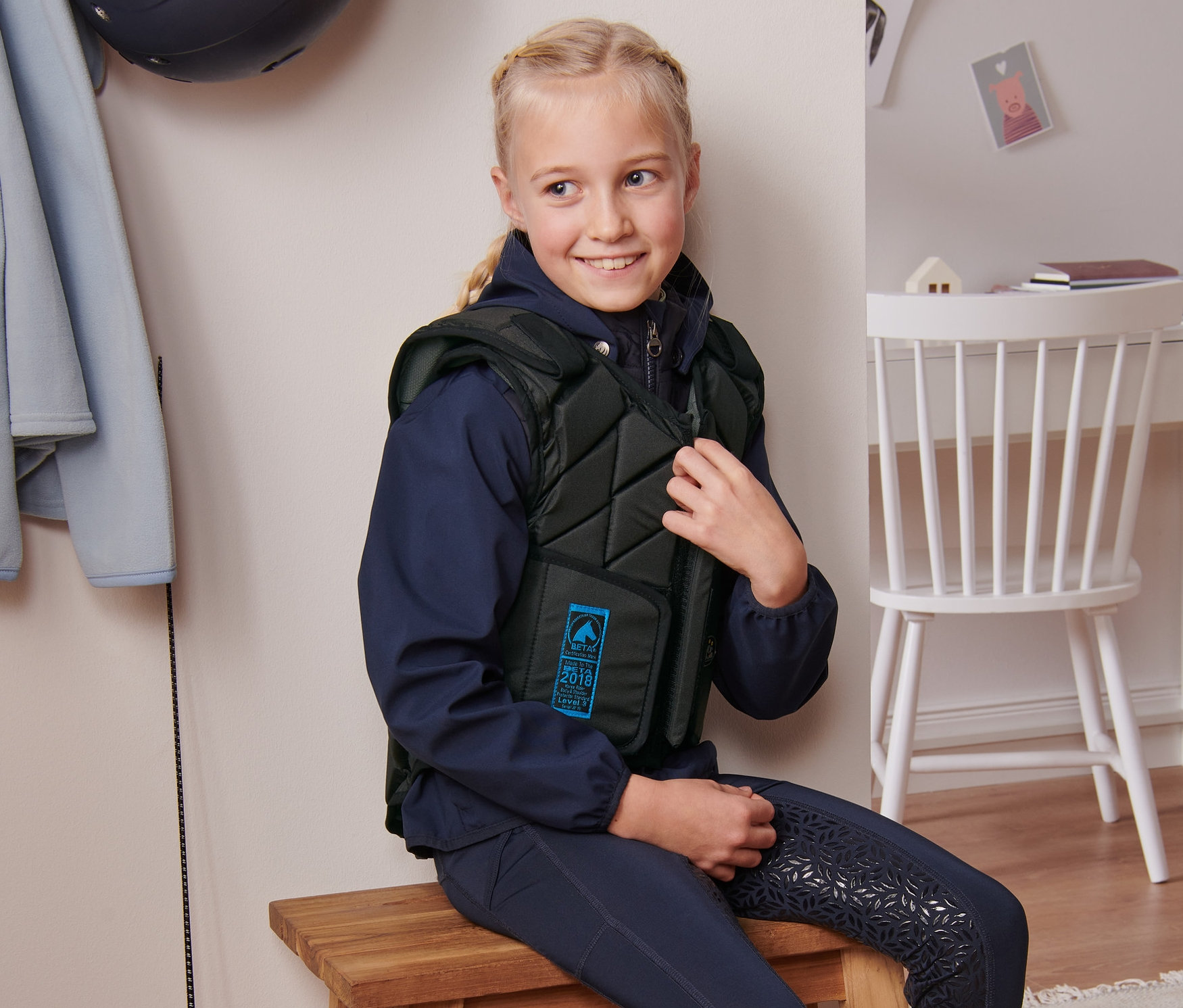 Mädchen sitzt auf einem Holzhocker und schliesst die USG Kinder-Reit-Sicherheitsweste »Eco Flexi«.