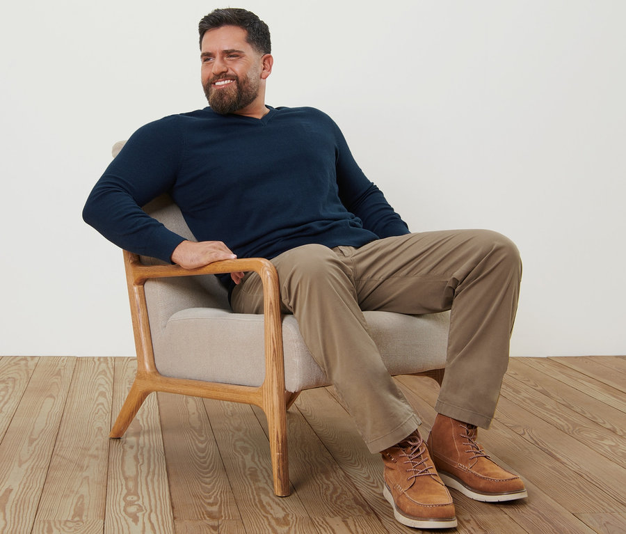 Ein Mann sitzt in einem Sessel, trägt sandfarbene Chinos und einen marineblauen Pullover mit V-Ausschnitt.