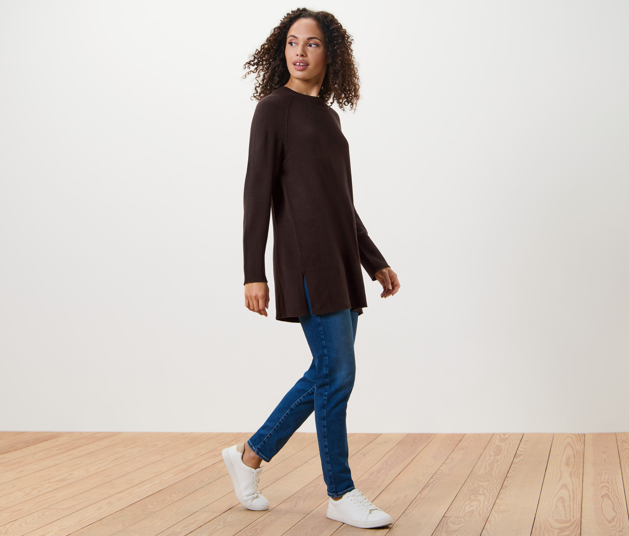 Frau geht und trägt einen langen Strickpullover in Dunkelbraun, Jeans und Turnschuhe.