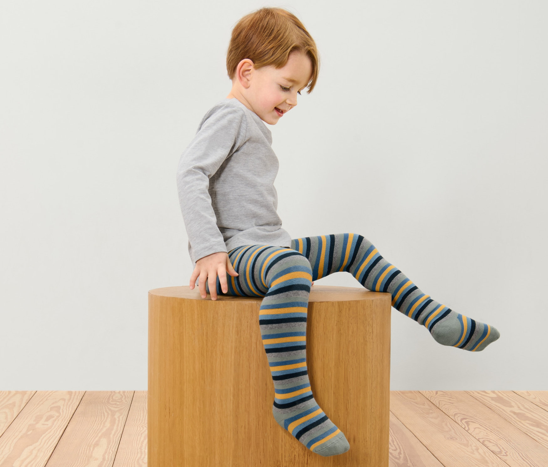 Ein Junge sitzt auf einem Holzhocker und trägt eine blaue, mehrfarbige Thermostrumpfhose.