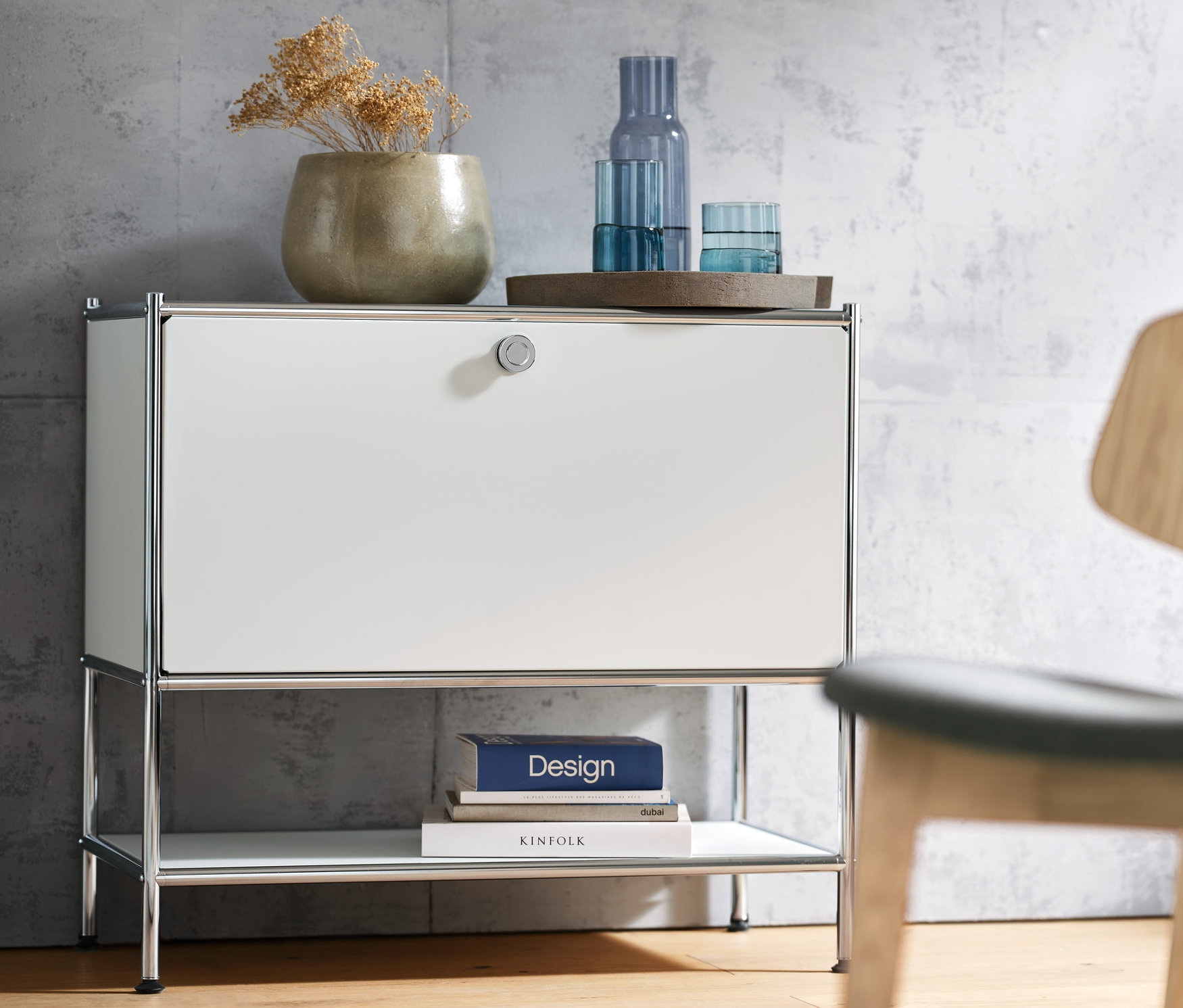 Weißes Sideboard »CN3« mit einer Klappe, darauf Vase und Gläser. Rechts ein Loungechair.