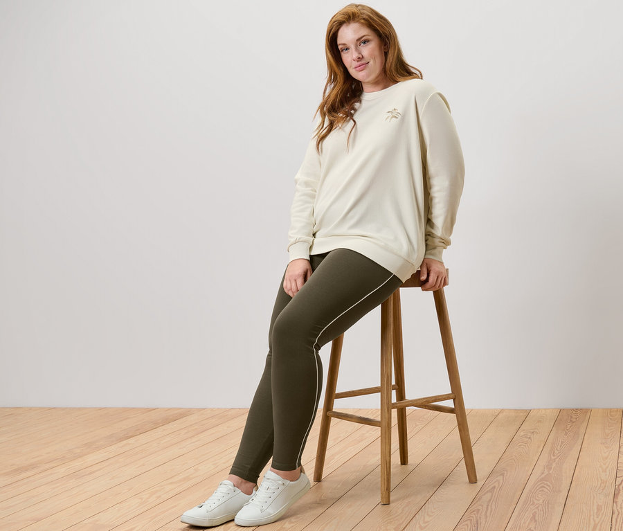 Frau sitzt auf einem Hocker, trägt ein Sweatshirt mit Stickerei und Leggings mit Paspelierung.