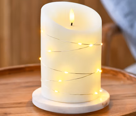 Eine LED-Echtwachskerze mit Microlichterkette steht auf einem runden Holztisch.