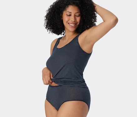 Lächelnde Frau mit lockigem Haar trägt ein dunkelblaues, gestreiftes SCHIESSER Tanktop und passende Midislips mit Modal. Sie zieht ihr Tanktop mit einer Hand hoch.