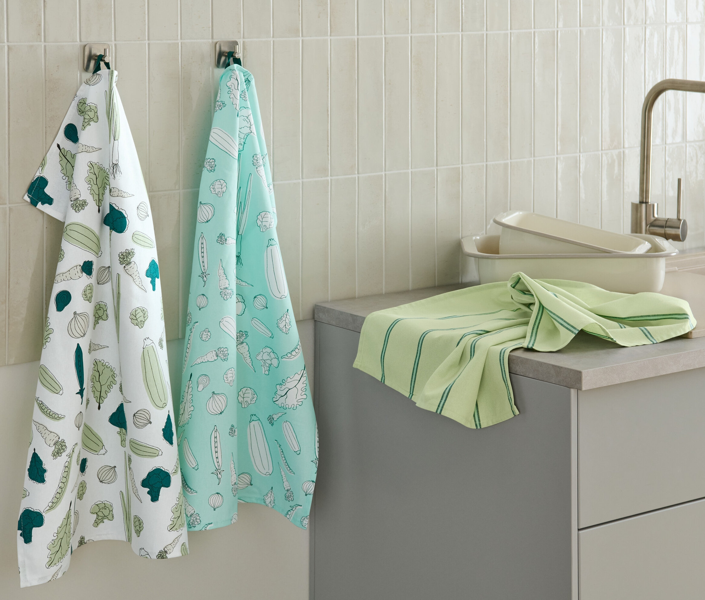 Zwei Geschirrtücher mit Gemüseaufdruck hängen an der Wand, ein weiteres grünes Geschirrtuch liegt auf der Küchentheke.