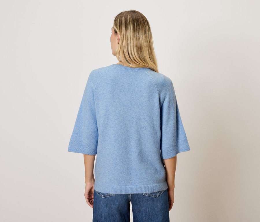 Frau mit blonden Haaren steht mit dem Rücken zur Kamera und trägt ein blaues Oberteil und Jeans.