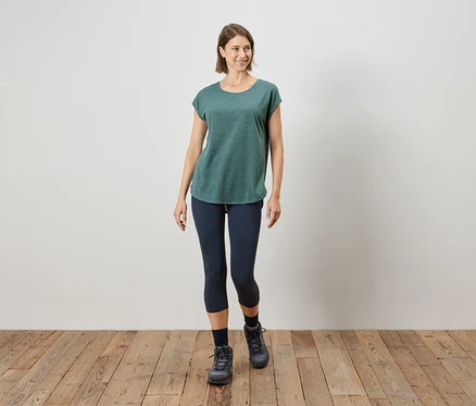 Frau geht in einem grünen T-Shirt, blauen Leggings und grauen Stiefeln auf einem Holzboden.