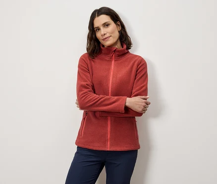 Frau posiert mit verschränkten Armen in orangefarbener Fleecejacke und marineblauer Hose.