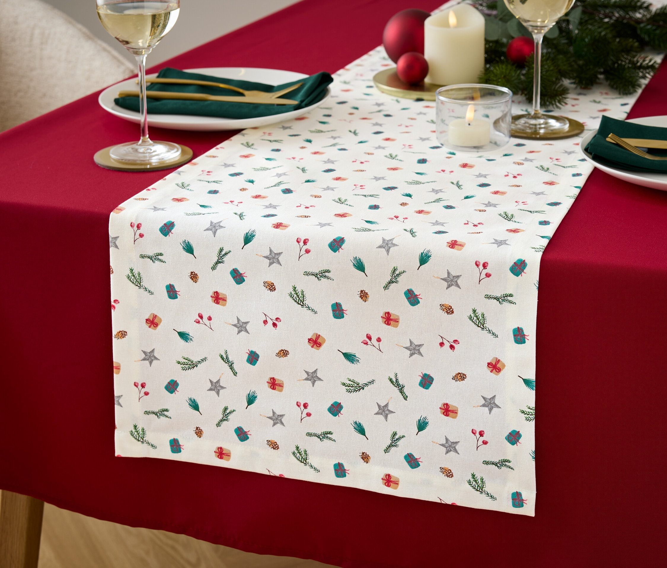 Gedeckter Tisch mit roter Tischdecke und weißem Tischläufer mit Stern-, Geschenk- und Zweigmuster.