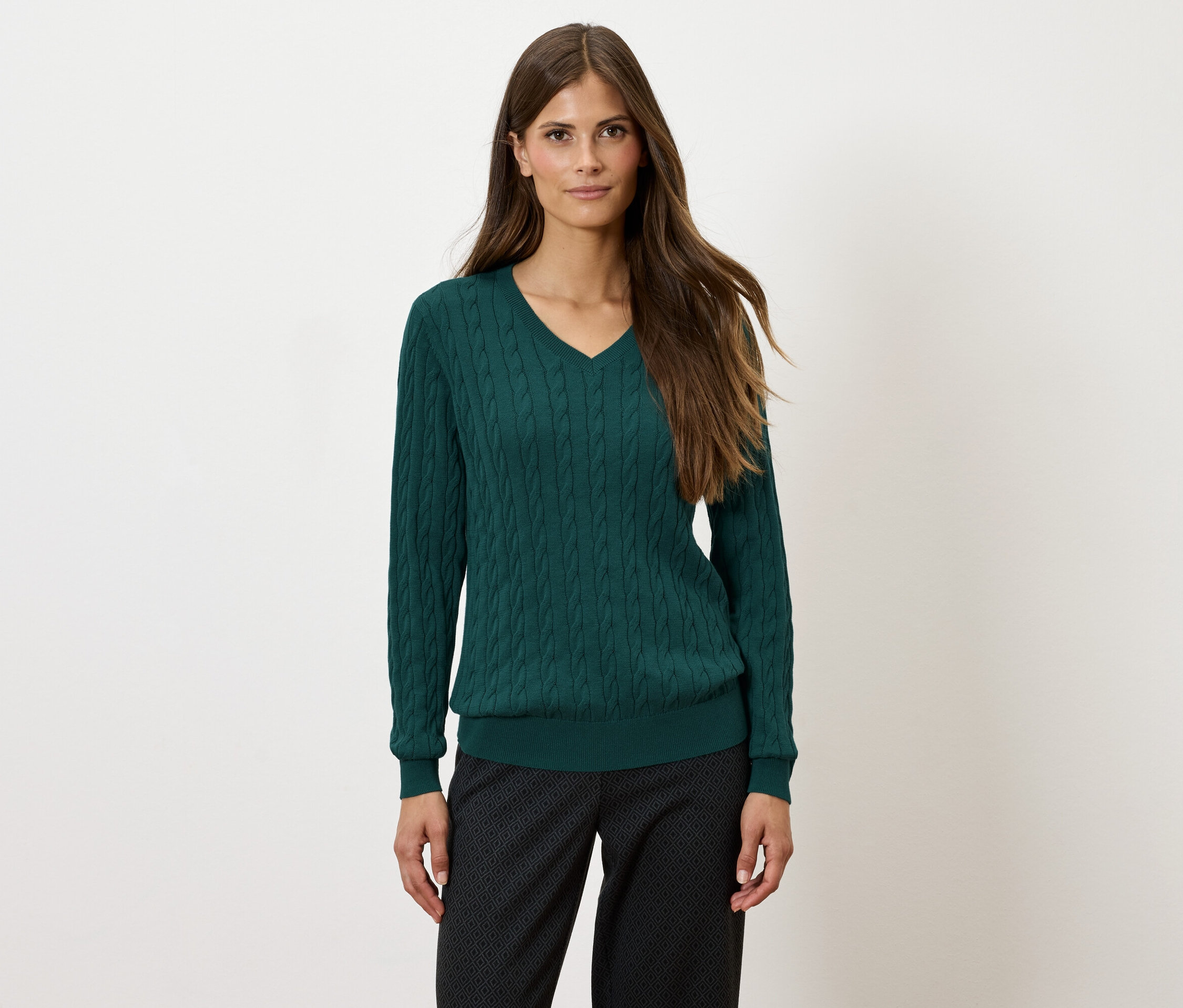 Frau mit langen braunen Haaren trägt einen grünen V-Ausschnittpullover und eine gemusterte, dunkle Hose.