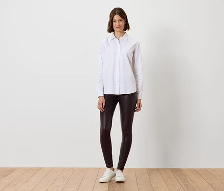 Frau im weißen Hemd und braunen Leggings steht auf Holzboden.