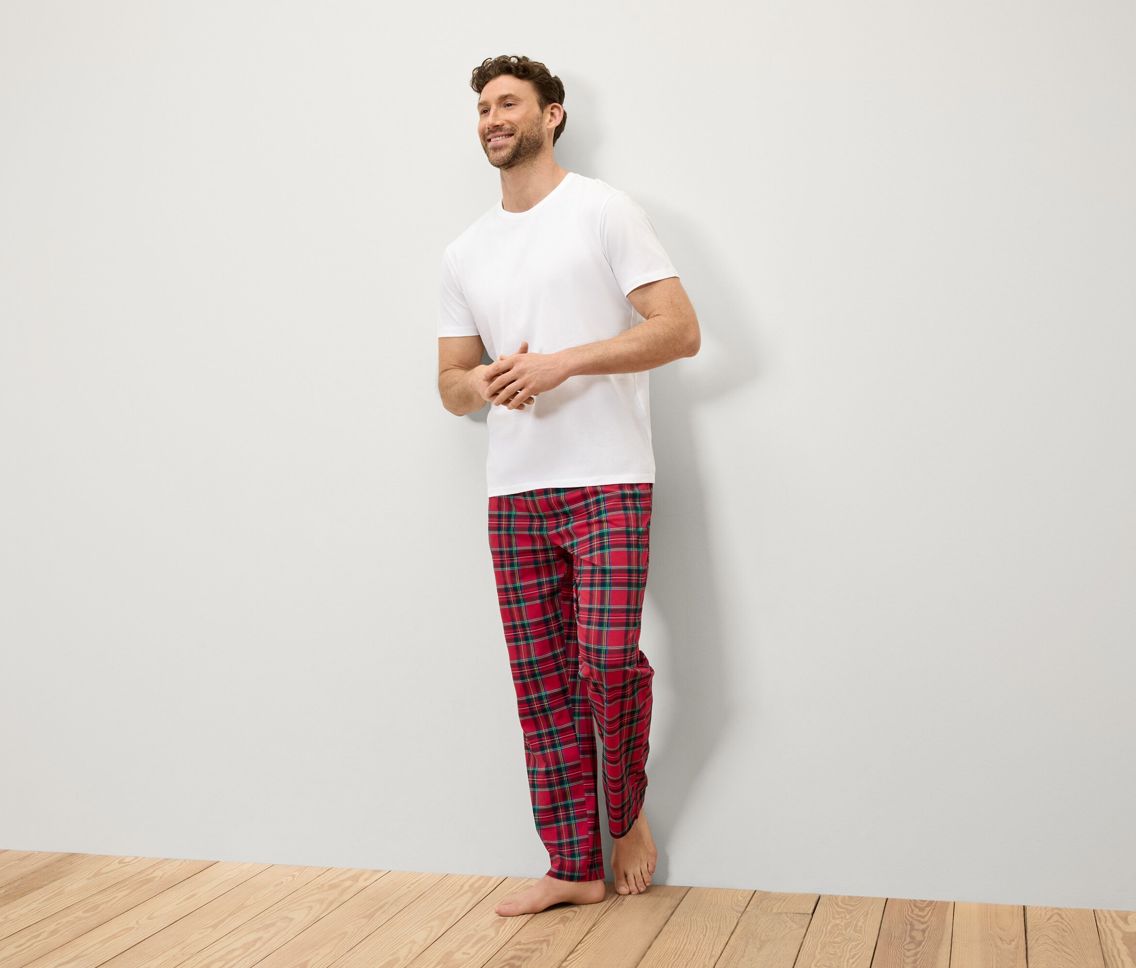 Ein lächelnder Mann steht an einer Wand und trägt ein weißes T-Shirt und eine rot karierte Hose.