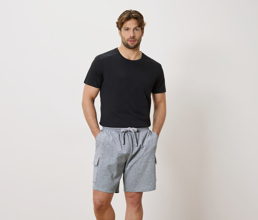 Mann mit Händen in den Taschen einer grauen Kordelzugshorts und einem schwarzen Kurzarm-T-Shirt.