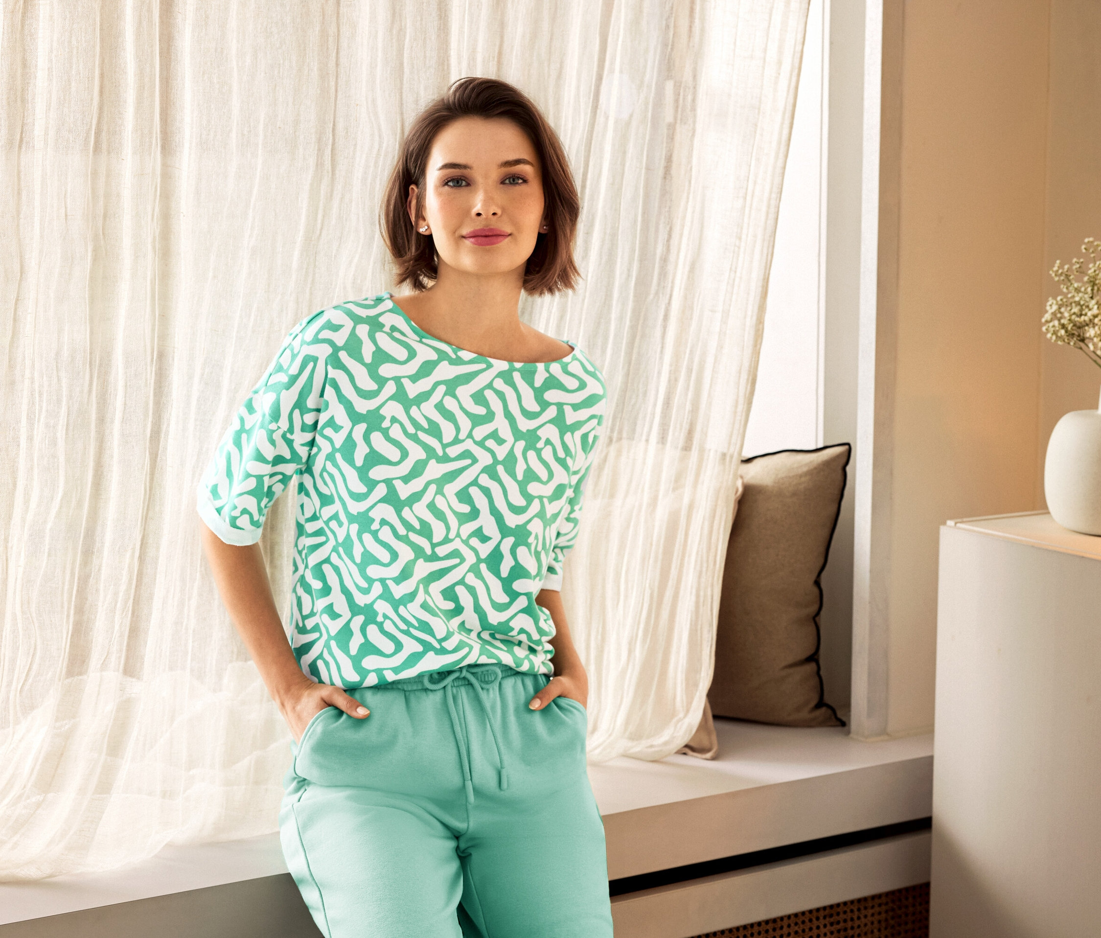 Frau mit grüner Hose und gemustertem Oberteil posiert neben einem Fenster. Im Hintergrund sind 925 Silber Ohrstecker Zirkoniabogen und eine Vase zu sehen.