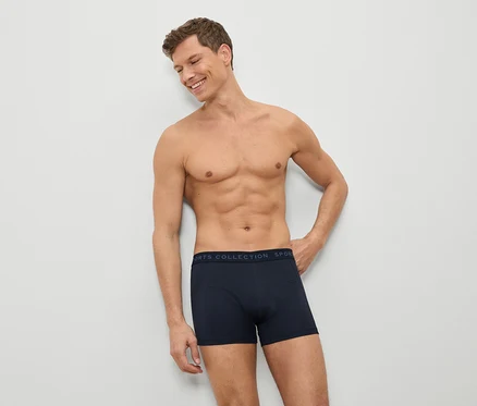 Ein Mann mit freiem Oberkörper und blauen Boxershorts lehnt an einer weißen Wand.