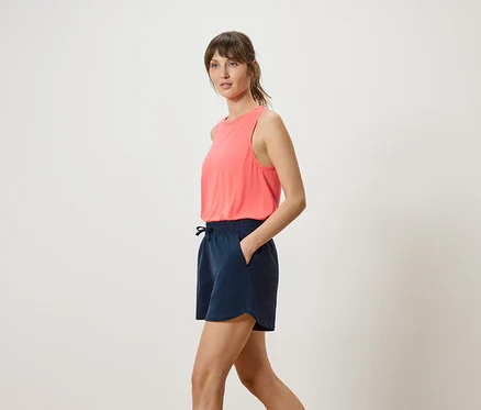 Frau mit korallfarbenem Tanktop und marineblauen Shorts, Hand in der Hosentasche, seitliche Ansicht.