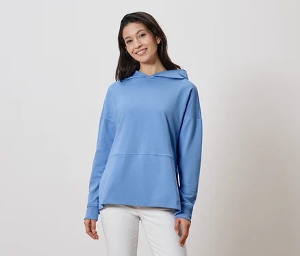 Frau mit langen, dunklen Haaren, die einen hellblauen Hoodie und weiße Hosen trägt.