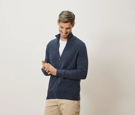 Ein Mann lächelt und reibt sich die Hände. Er trägt eine marineblaue Strickjacke und eine beige Hose.