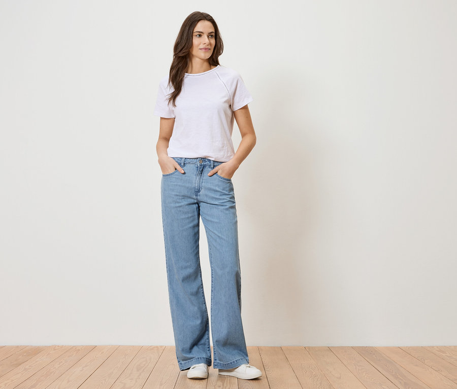 Frau mit weißem T-Shirt und blauen Jeans steht vor einer weißen Wand.