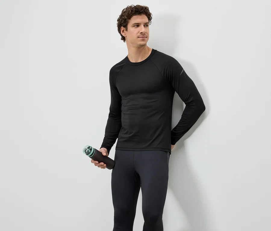 Mann in schwarzem Sportoberteil und Leggings hält eine braune Wasserflasche vor einer weißen Wand.