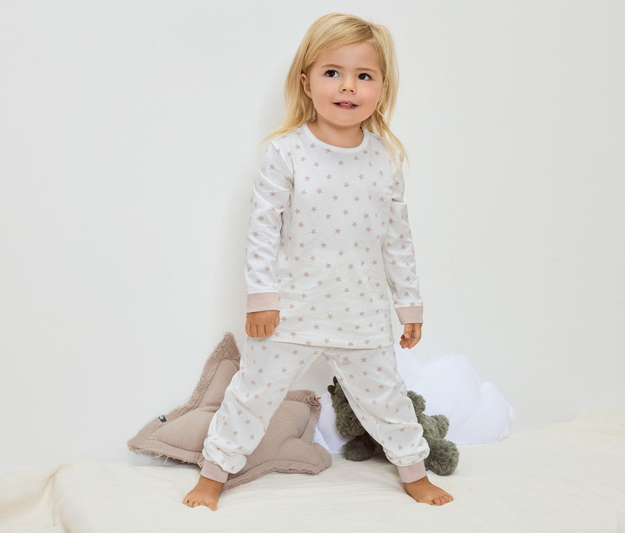 Ein kleines blondes Mädchen steht barfuß auf einer Decke und trägt einen weißen Schlafanzug mit Sternenmuster.