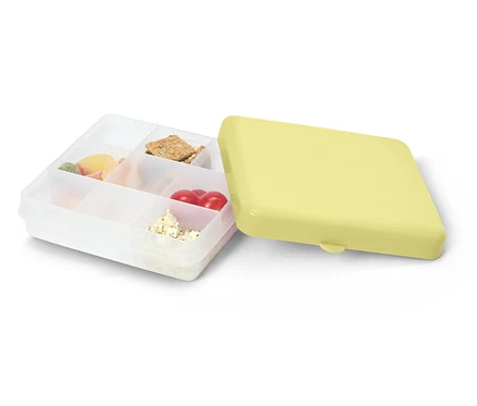 Geöffnete, durchsichtige Lunchbox mit gelbem Deckel. Gefüllt mit Süßigkeiten, Crackern, Tomaten und Käse.