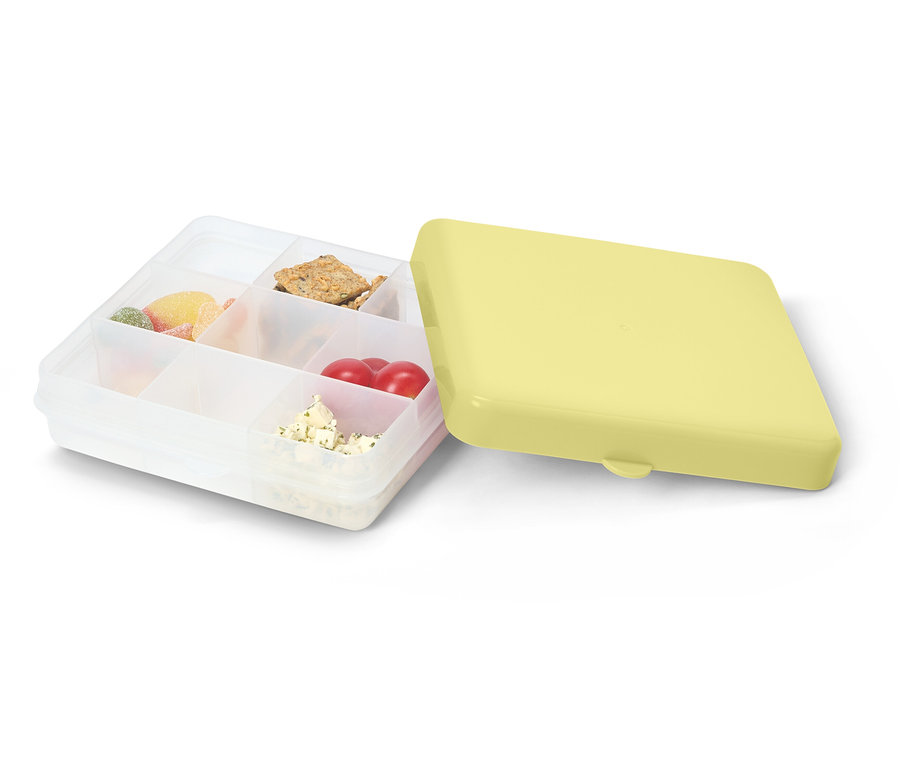 Geöffnete, durchsichtige Lunchbox mit gelbem Deckel. Gefüllt mit Süßigkeiten, Crackern, Tomaten und Käse.