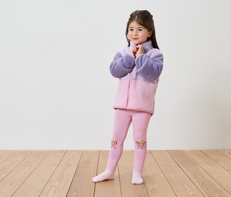 Ein Mädchen in einer lila-rosa Fleecejacke und rosa Leggings mit Einhorn-Motiv steht auf einem Holzboden vor einer weißen Wand.