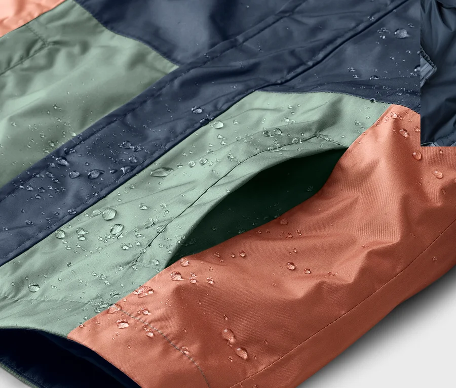 Nahaufnahme einer wasserabweisenden, farblich abgesetzten Jacke in Blau, Grün und Orange mit Wassertropfen und einer Tasche.