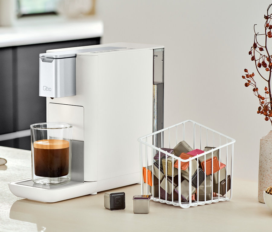 Eine Qbo ESSENTIAL Bright Sand Kaffeemaschine steht neben einem Glas Kaffee auf einer Küchentheke.