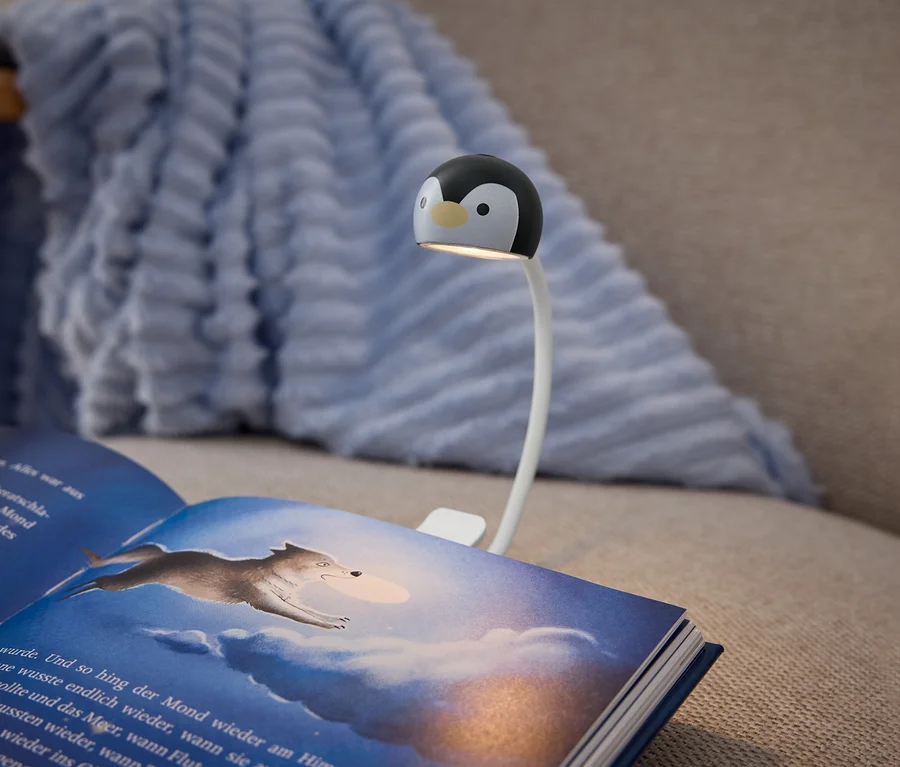 Ein aufgeschlagenes Buch mit dunkelblauem Einband liegt auf einem beigen Sofa, beleuchtet von einer Klemmleuchte. Die Leuchte hat einen weißen, flexiblen Arm und einen schwarz-weißen Pinguinkopf, der Licht auf die Buchseiten wirft. Die rechte Seite zeigt eine Illustration eines Wolfs, der durch einen bewölkten, blauen Himmel springt. Eine hellblaue, strukturierte Decke liegt im Hintergrund über der Sofalehne.