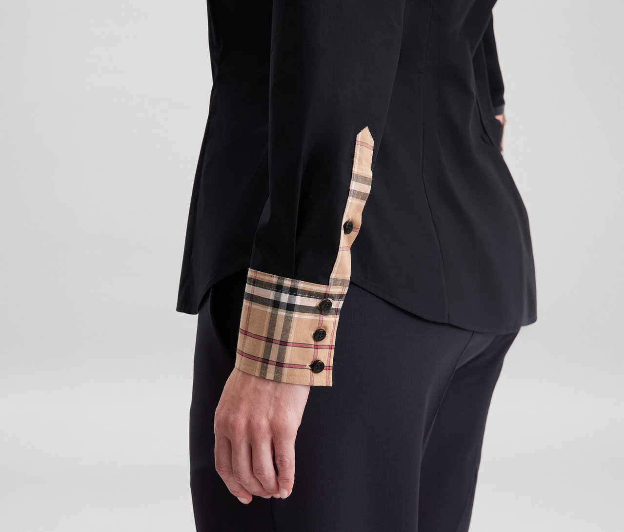 Nahaufnahme einer Person in einer schwarzen Seidensticker Popeline-Bluse mit beigen, schwarzen und roten Karomustermanschetten und schwarzen Knöpfen, kombiniert mit einer dunklen Hose.