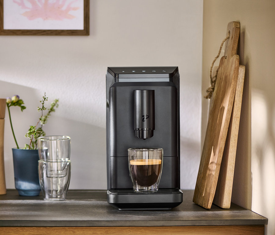 Grauer »Esperto mini« Kaffeevollautomat füllt Kaffee in ein Glas, daneben Schneidebretter, Gewürzmühlen und Gläser auf einer Arbeitsplatte.