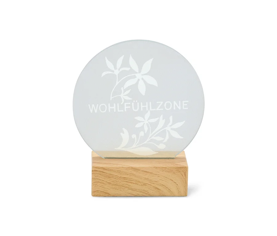 Rundes, durchscheinendes Schild mit der Aufschrift "WOHLFÜHLZONE" und Blumenmustern auf einem hellen Holzsockel.
