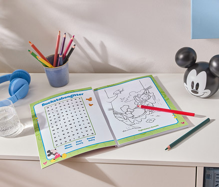 Auf einem weißen Tisch liegen ein aufgeschlagenes Malbuch, bunte Stifte in einem blauen Becher, blaue Kopfhörer, ein Glas Wasser und eine Mickey Mouse-Figur.