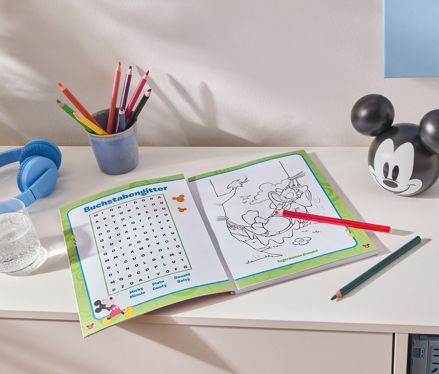 Auf einem weißen Tisch liegen ein aufgeschlagenes Malbuch, bunte Stifte in einem blauen Becher, blaue Kopfhörer, ein Glas Wasser und eine Mickey Mouse-Figur.