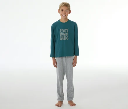 Ein lächelnder Junge steht barfuß und trägt einen SCHIESSER Kinder-Pyjama »Skate« mit einem dunkelgrünen Langarmshirt, auf dem "SKATE BOARDING" steht, und einer grauen Hose.