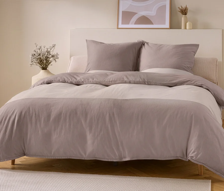 Ein Bett mit hellem Kopfteil ist mit hellbraun-taupe Flanell-Bettwäsche bezogen, die horizontale Streifen in zwei Farbtönen aufweist. Zwei taupefarbene Kissen liegen am Kopfteil. Links steht eine Vase mit Trockenblumen, rechts ein Holzhocker mit Buch.