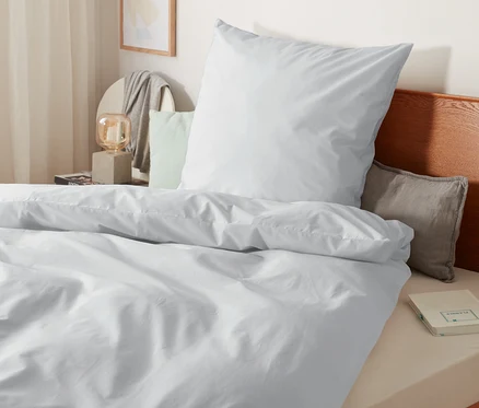 Detailansicht von weißer Perkal-Bettwäsche in Normalgröße, Übergröße und Doppelgröße auf einem Bett mit Holzrahmen.