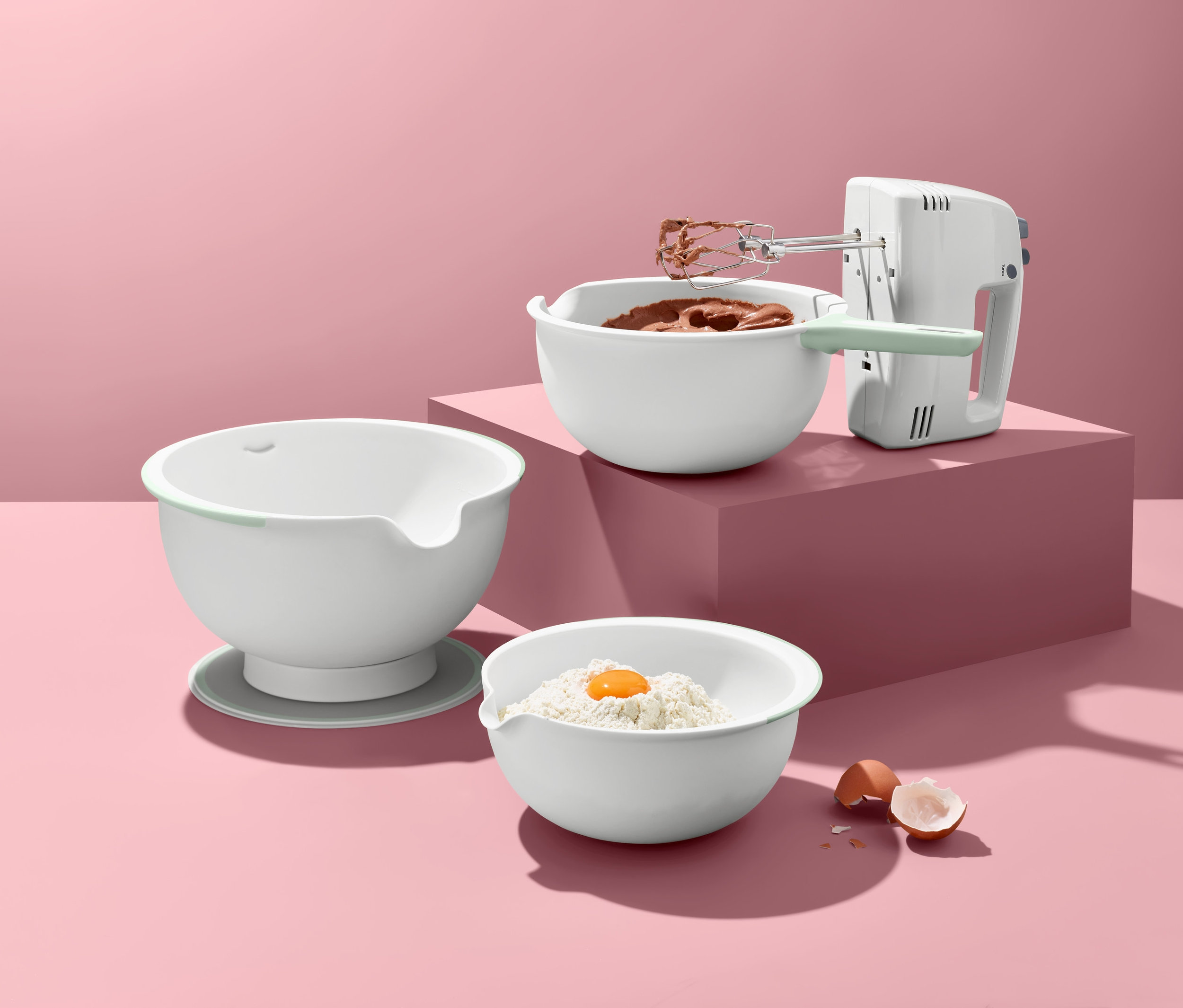 Ein Mixer steht neben einer Schüssel mit Schokoladenmischung; darunter liegt eine Schüssel mit Mehl und Ei; daneben liegt eine leere Schale; und dahinter ein Rührschüssel-Set.