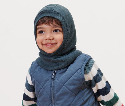 Lächelnder Junge trägt eine Kleinkind-Schlupfmütze, eine Steppweste und einen gestreiften Pullover.