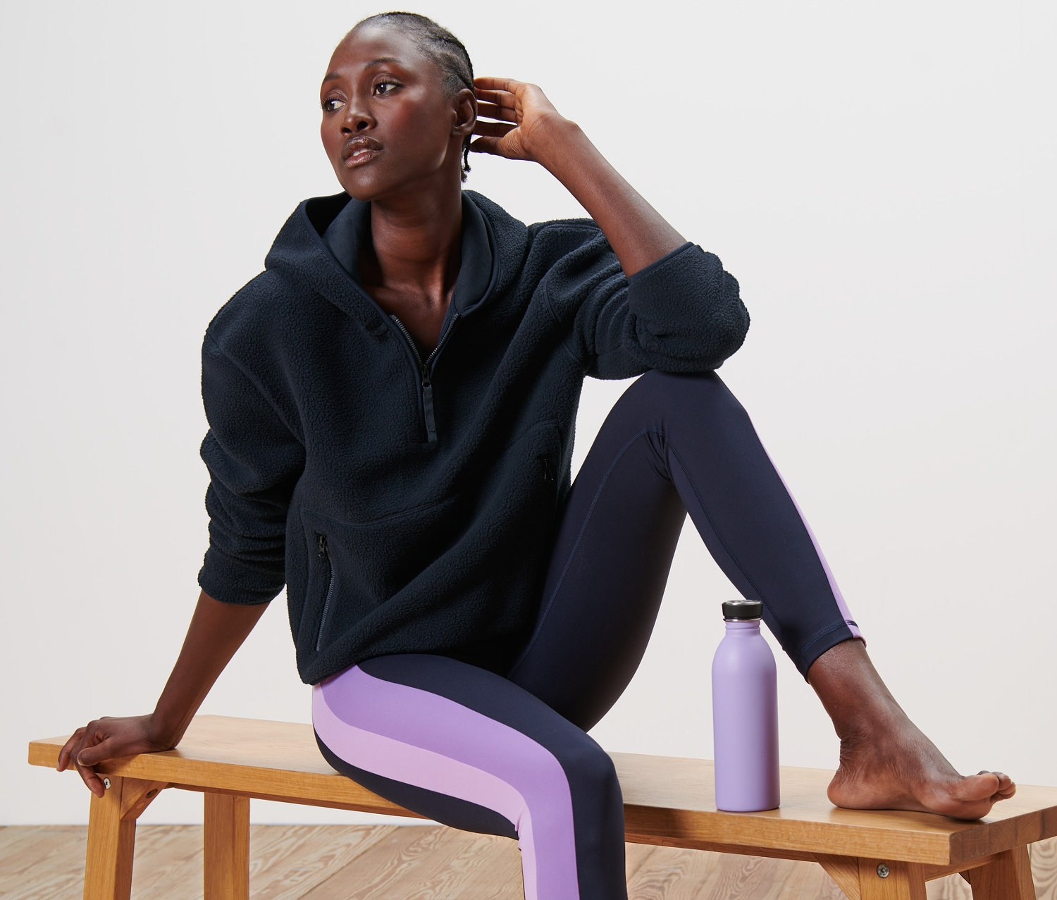 Frau sitzt auf einer Bank in einem dunkelblauen Teddy-Kapuzenpullover und Leggings mit lila Streifen. Neben ihr steht eine lila Wasserflasche.