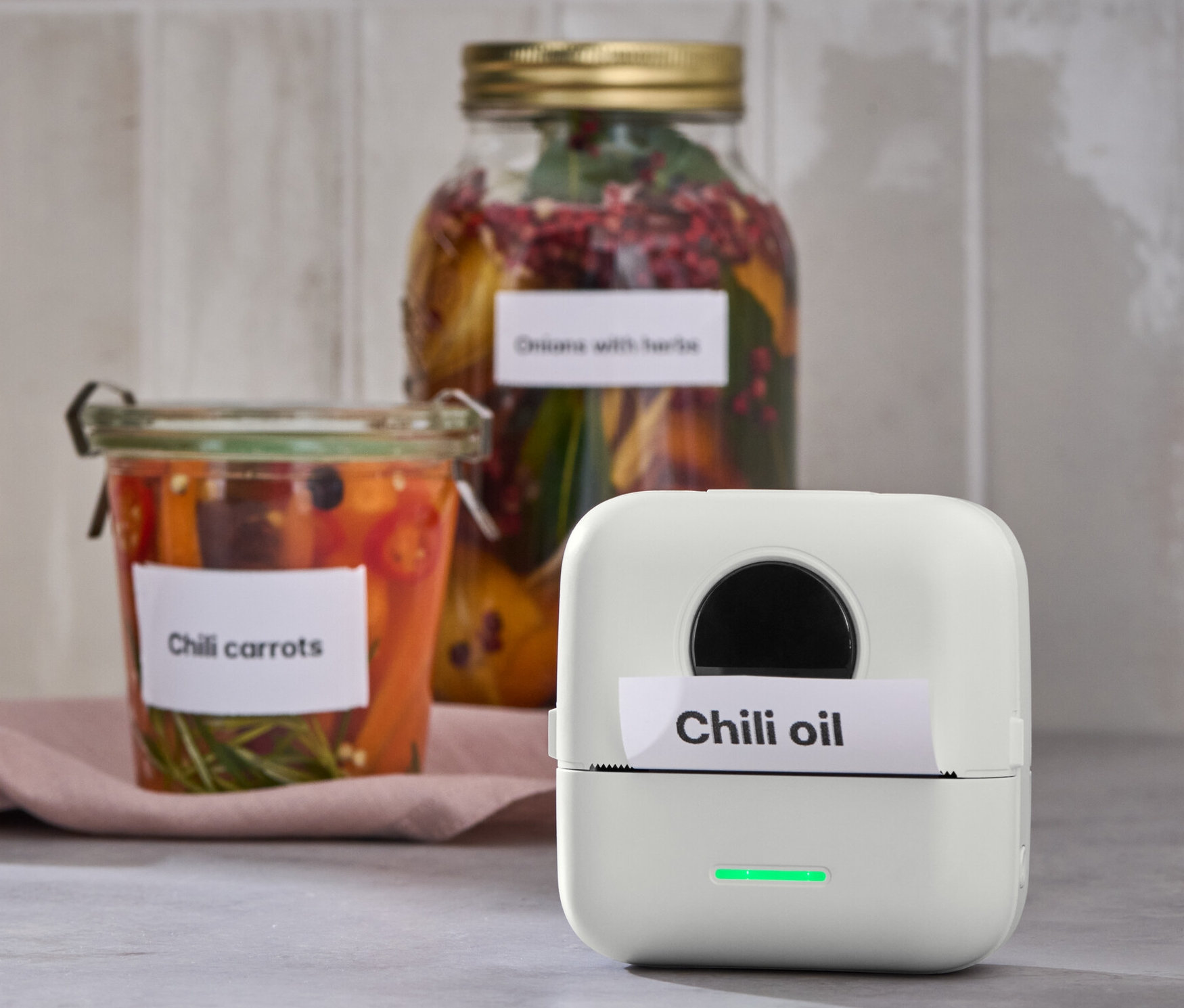 Ein tragbarer Mini-Thermodrucker druckt ein Etikett mit der Aufschrift Chiliöl. Im Hintergrund Gläser mit Chili-Karotten.