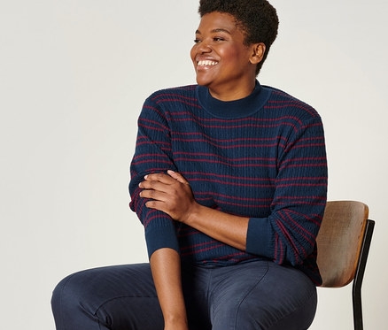 Eine Frau sitzt auf einem Stuhl und trägt einen blau-roten Feinstrickpullover mit Zopfmuster, eine dunkelblaue Twillhose mit Komfortbund und Leder-Loafer.