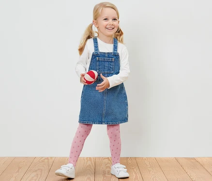 Ein lächelndes blondes Mädchen in einem Jeanskleid hält einen rot-weissen Ball und trägt 2 Kinder-Feinstrumpfhosen.