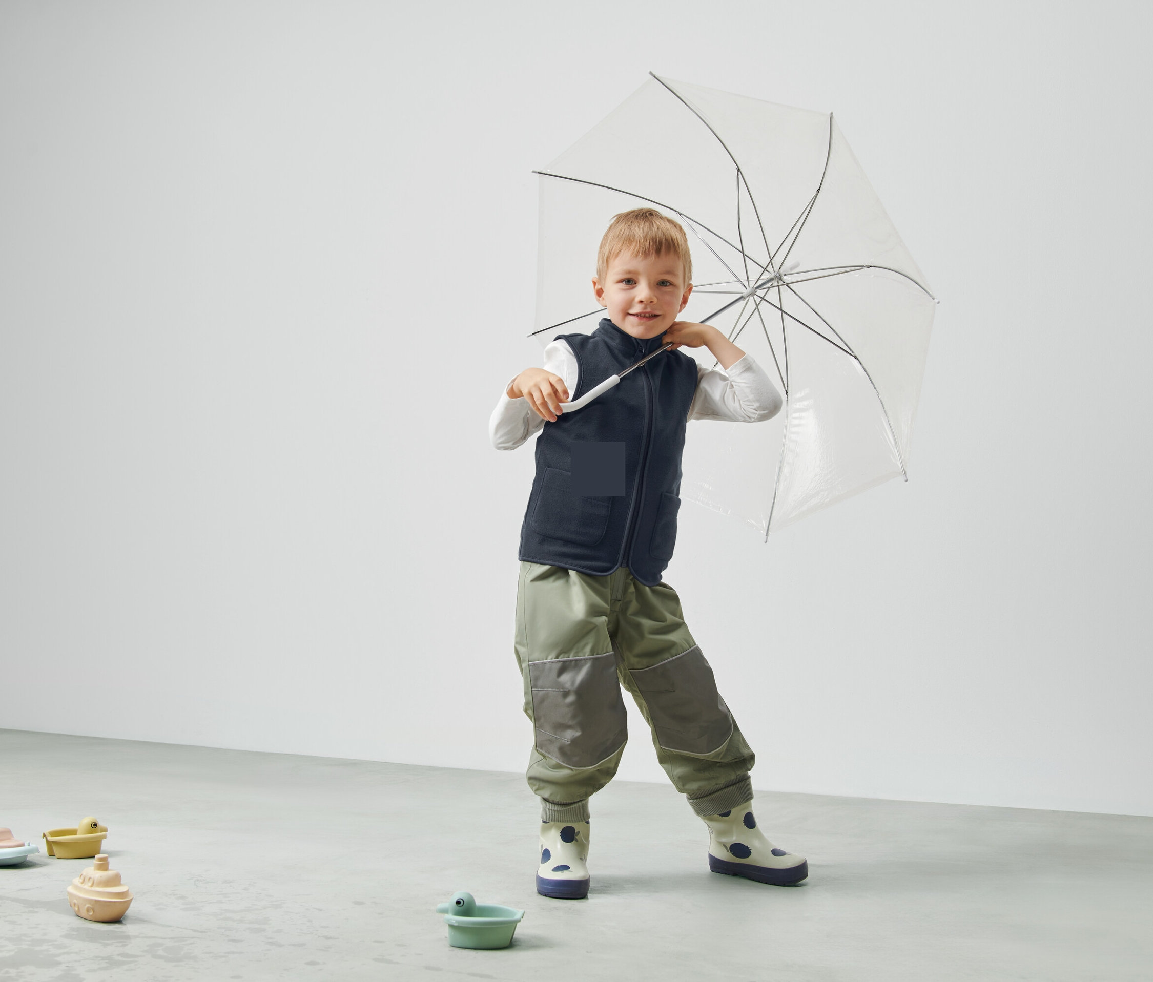 Junge hält einen durchsichtigen Regenschirm, trägt eine Weste und Hose und steht in einem Raum.