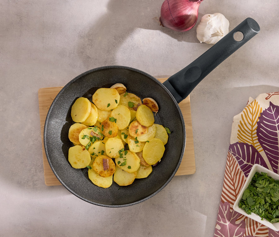 Bratkartoffeln in einer schwarzen Pfanne auf einem Holzbrett mit Knoblauch, Zwiebeln und Petersilie.