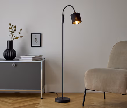 Eine schwarze Stehlampe steht neben einer grauen Kommode, auf der eine Vase und Bücher stehen. Neben der Kommode steht ein beiger Sessel.