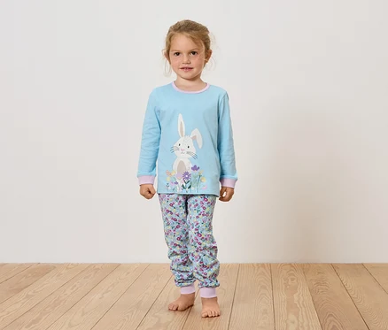 Mädchen in hellblauem Pyjama mit Hasen- und Blumendruck steht auf Holzboden.