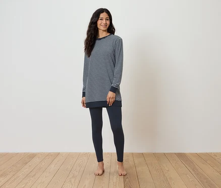 Eine barfuß stehende Frau auf einem Holzboden trägt ein gestreiftes Langarmshirt und dunkelblaue Leggings.
